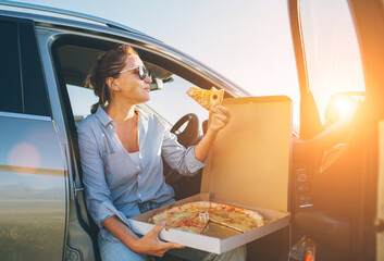 Midle-aged woman eating just cooked italian pizza sitting on driver car seat during meal break and...
