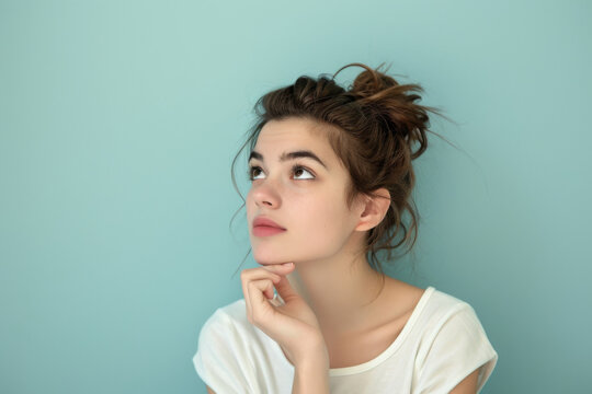 Contemplative thinking young female with a messy bun looking upwards with a thoughtful expression, isolated on a pale blue studio background