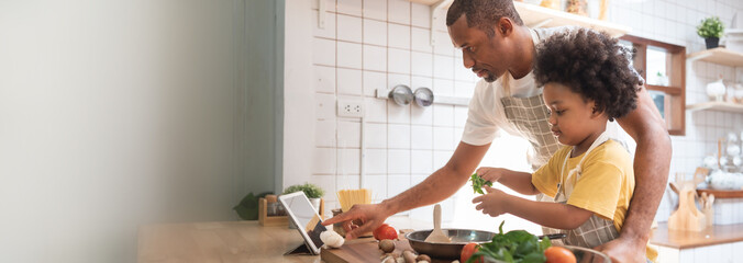 African American Little boy preparing food while his father looking on the digital recipe and using...