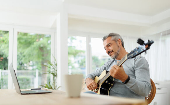 Mature man enjoying a guitar session in a white room with a laptop and coffee mug on the table.