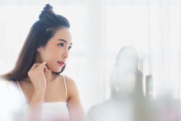 Pretty Asian Korean woman model looking at make-up mirror and touch her face at home. Skin care,...