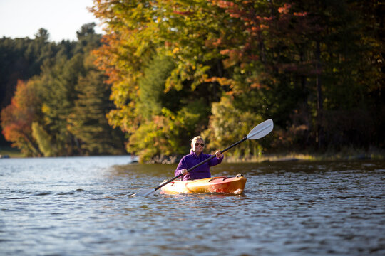 Woman kayaking with autumn foliage on pond or lake with blonde hair and orange boat and purple jacket in Vermont and New England.