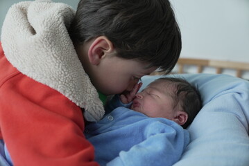 A touching moment as an older child in a red jacket gently leans in to nuzzle their newborn sibling, who is wrapped in a blue onesie. The intimate moment captures the bond between siblings.