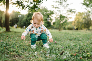 Boy catches soap bubbles in nature. Toddler little cute walking and playing in spring garden. 2 year old boy enjoying on green grass in park on sunny summer day. Outdoors creative activities for kids.