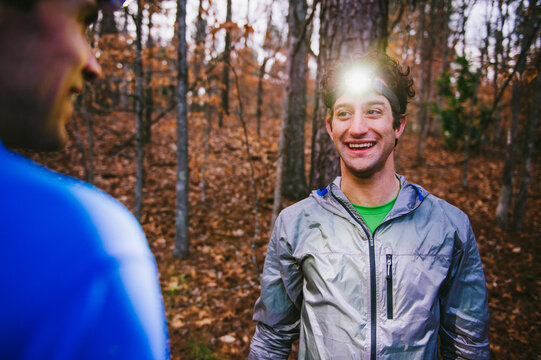 Two young men wearing headlamps and hiking clothes smile at each other in the woods in the evening