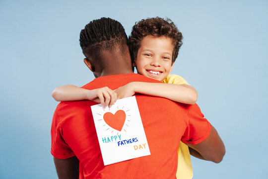 Cute African American boy holding father's day greeting card, hugging his dad, looking at camera
