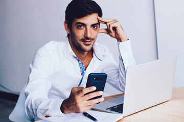 Portrait of handsome male director working online creating presentation via laptop application and looking at camera during break, indian entrepreneur chatting with colleagues on modern smartphone