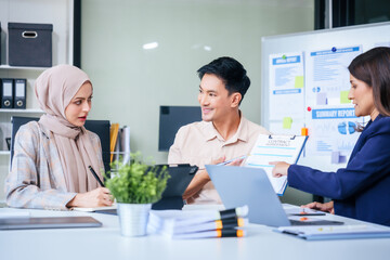 A diverse group of professionals, including a male Asian businessman and a female Muslim businesswoman in hijab, gather around a desk equipped with tablets and laptops for a productive meeting.