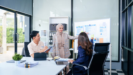 A multicultural team of professionals, including male and female businesspersons, convenes around a desk with tablets, discussing strategies illustrated on a whiteboard during a dynamic presentation.