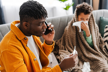 Sick African American father and little son have cold, man talking to doctor, measuring temperature