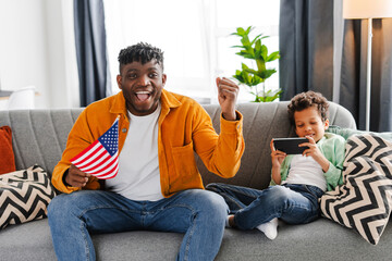 Smiling African American father and little son sitting on sofa at home, man holding American flag