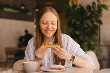 Young blonde woman eating eclair sitting in cafe. Girl breaks the eclair look joyful wow at...
