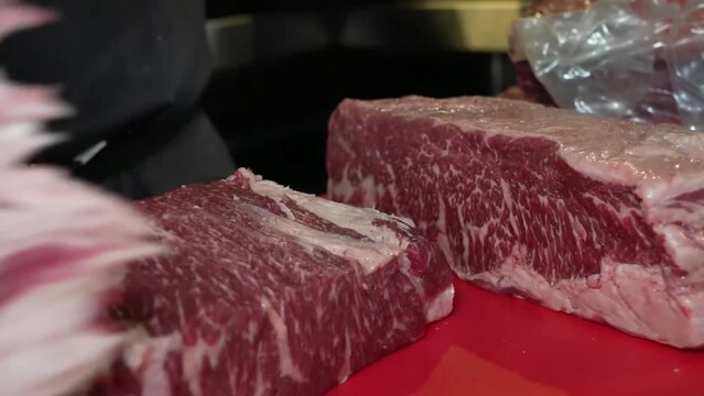 Person, wearing white gloves, skillfully trimming excess fat, and veins from a piece of marbled meat using a knife on a red cutting board.