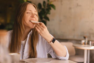 Young blonde woman eating eclair sitting in cafe. Girl bite piece of croissant look joyful at...