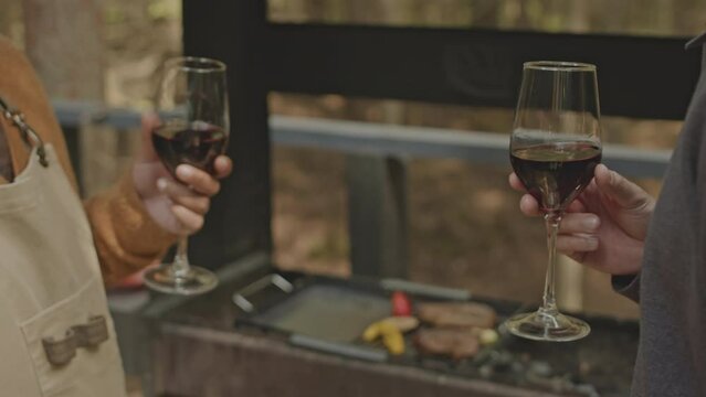Close up view of hands of two male friend clinking glasses with red wine in toast while having party on outdoor terrace beside BBQ grill