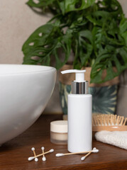 White one pump bottle near basin and green plant on wooden countertop in bath, mockup