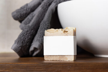 Soap bar with blank label near grey towel on basin on wooden countertop in bath, mockup