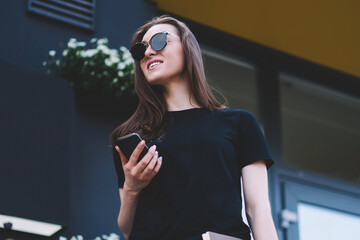 Positive female student in sunglasses holding smartphone device trying connect to wireless 4g...
