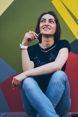 Fototapeta premium Pondering positive woman on colorful vivid background dreaming about vacations with friends, happy thoughtful hipster girl holding spectacles and enjoying free time on city streets near bright wall