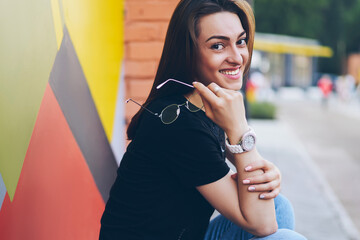 Portrait of cheerful female student dressed in denim design apparel sitting on urban setting and...
