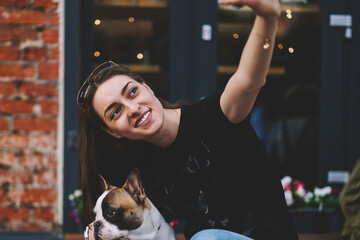 Happy smiling hipster girl clicking selfie photo on modern smartphone web camera sitting on bench with french bulldog, positive woman posing with animal friend and taking images for own blog about pet