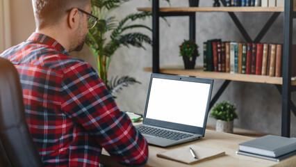 Back view of business man freelancer sitting in home office at table,using laptop with white screen...