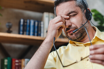 Close up of call center operator wearing headset tired at home office. Handsome middle age man...