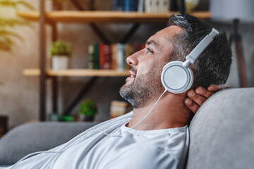 Middle aged bearded man listening music with headphones relaxed in sofa at his home copy space,...