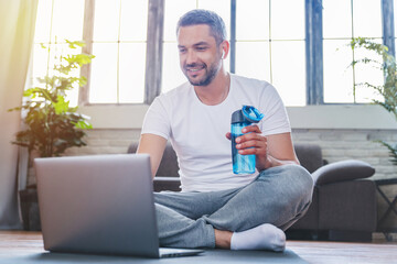 Happy mature caucasian man in sportswear sitting on yoga mat with bottle of water, choosing fitness...
