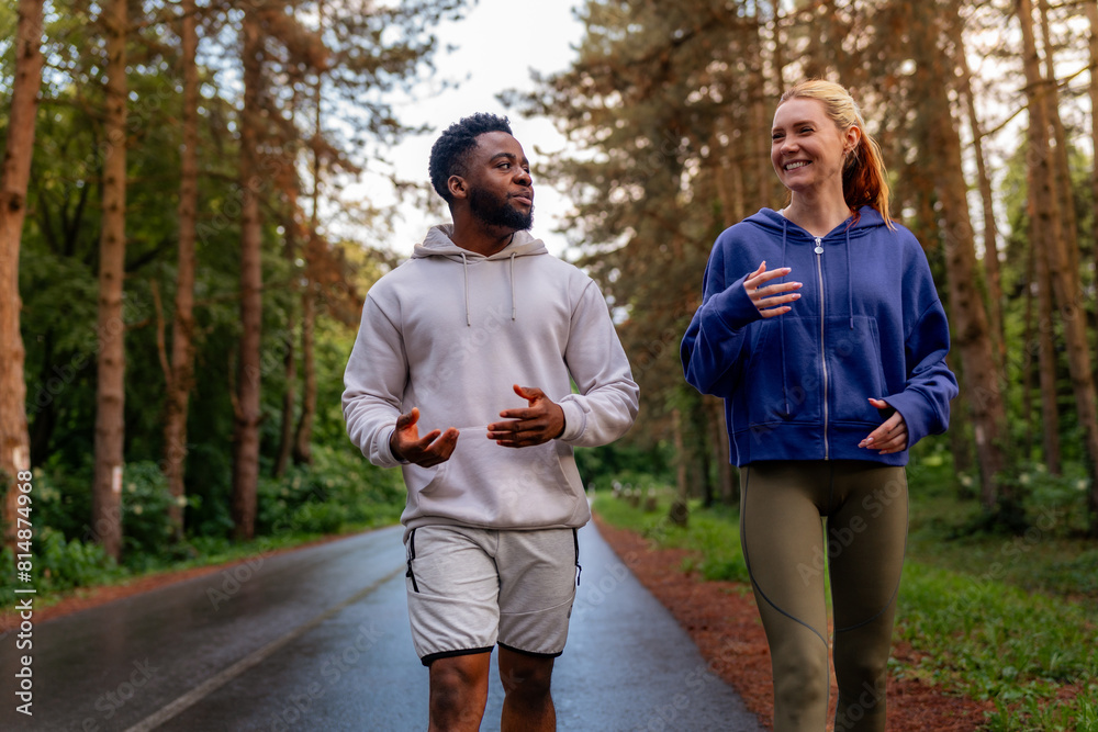 Wall mural Two friends running through a scenic forest path, sharing joy and fitness. Wearing hoodies and athletic gear, they highlight the importance of exercise and companionship. - Wall murals