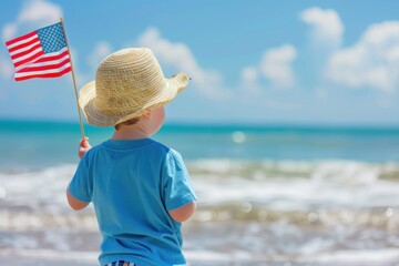 A young Caucasian boy in a blue shirt and straw hat holds an American flag at the beach. Memorial day, independence day
