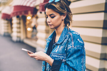Serious hipster girl in trendy jeans jacket typing mail and reading news on website via cellphone,...