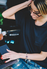 Trendy clever hipster girl wear in spectacles for provide eyes protection working via computer while spending time at street cafe, concentrated female programmer thinking about creating new web site