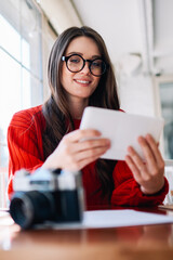 Cheerful young woman received positive message on gadget checking notification, happy skilled hipster girl in eyeglasses reading news via touchpad at cafeteria and writing funny article for sharing