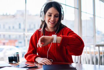 Cheerful hipster girl listening new bestseller audiobook using application and earphones sitting at coffee shop, positive teenager enjoying funny playlist and checking time on wearable smartwatch