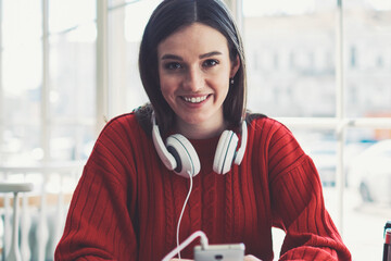 Portrait of young cheerful hipster girl looking at camera holding cellular and choosing music for...