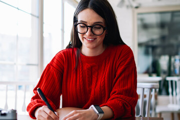 Smiling positive female student journalist sitting at table and writing essay exited with idea, successful hipster girl in eyeglasses enjoying time at university campus while completing homework
