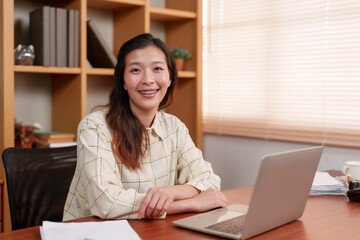Smiling Asian woman sitting at desk in bright office. Wearing checkered shirt, looking at camera, with laptop, documents, and wooden shelves in background. Professional, confident, and approachable.