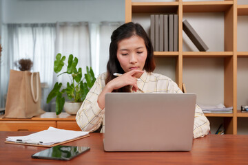 Focused Asian woman in checkered shirt sitting at desk working on laptop in bright office. Holding...