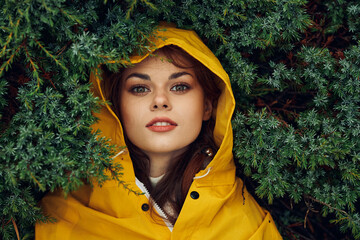 A young woman in a yellow raincoat hiding behind green leaves, exploring nature on a rainy day