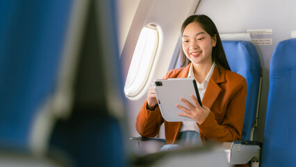 Asian woman holding tablet while sitting on airplane near window during flight, travel and business...