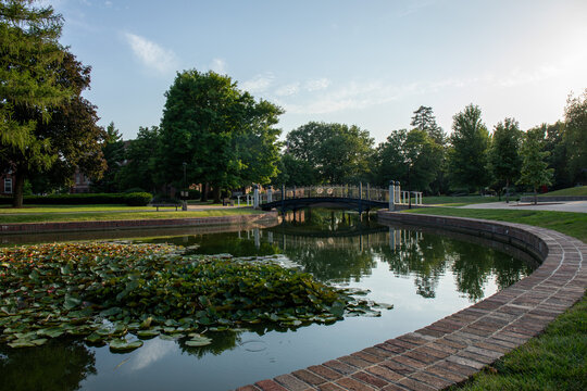 Green trees and pond with reflective surface.  Pedestrian bridge over clear smooth pond.  Central College campus in Pella, Iowa.  