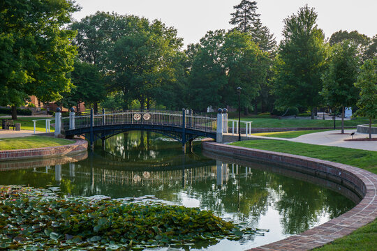 Green trees and pond with reflective surface.  Pedestrian bridge over clear smooth pond.  Central College campus in Pella, Iowa.  