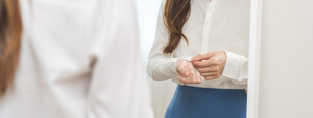 Get dress, asian young woman, businesswoman standing, hand buttoning up shirt formal, female getting dressed, ready before go to work looking reflection the mirror in bedroom in the morning at home.