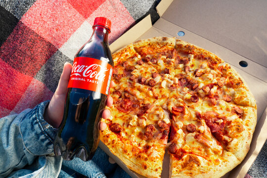 Krivoy Rog, Ukraine, May 1, 2024: A bottle of Coca-Cola and a box of pizza in hands against the background of a plaid on green grass. Outdoor picnic. View from above. Close-up