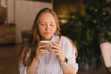 Portrait of gorgeous blonde smiling lady smelling, enjoying of coffee and drinking cappuccino from cup while resting in restaurant. Woman wear stripped shirt.