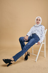 Fototapeta premium Portrait of an Asian model in white shirt and blue jeans with hijab sitting on a white chair isolated over beige background. Stylish Muslim female hijab fashion lifestyle concept.