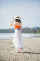 A woman in a hat stands with her back walking on the beach,A girl in a long white dress and hat is walking along the coast with a book in her hand.