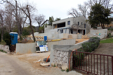 Construction site for modern villa by the sea filled with construction material mixed with wooden supports and glass windows waiting to be installed surrounded with trees without leaves and portable