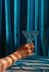 Female hand holding a martini glass against the background of a blue curtain. Modern art.